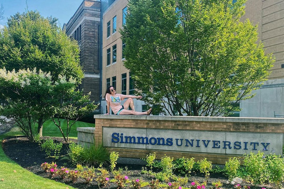 Undergraduate Simmons student sitting on the Simmons University sign in Boston, Massachusetts.