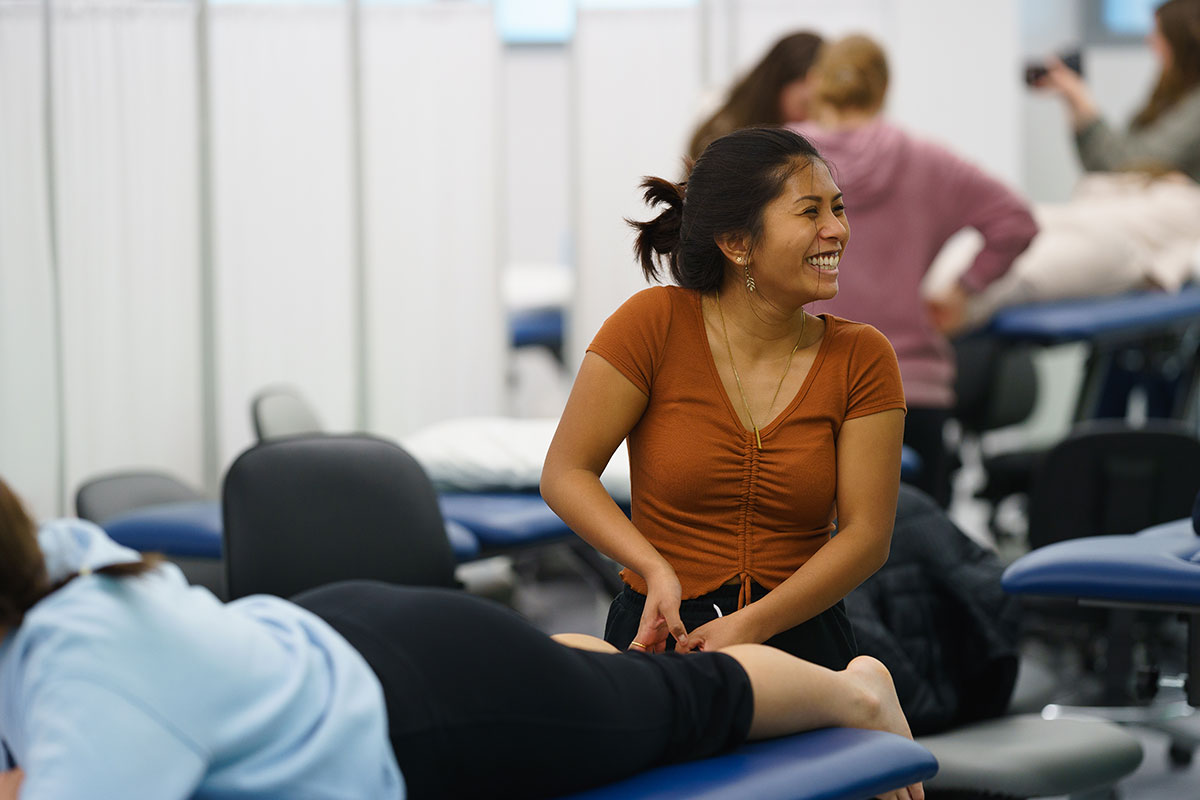 A student in the Simmons University Physical Therapy lab working with a mock patient and smiling