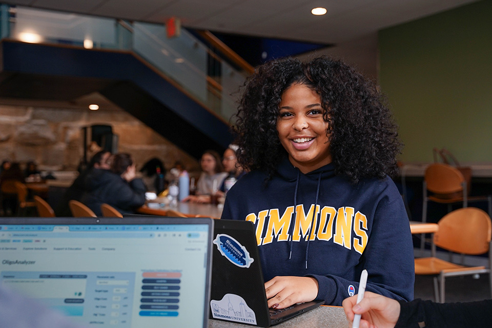 Student in Simmons sweatshirt smiling while on laptop in the Fens