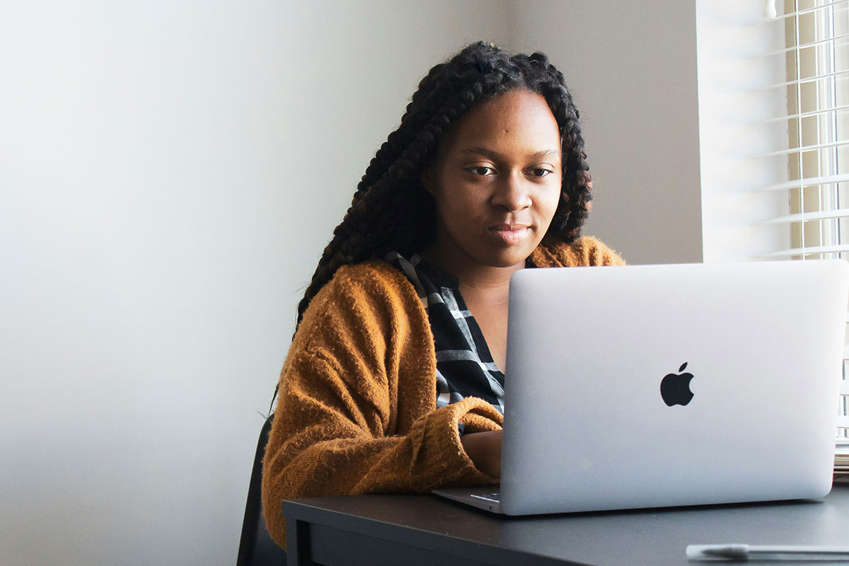 A young woman wearing an orange sweater while working on a laptop near a window