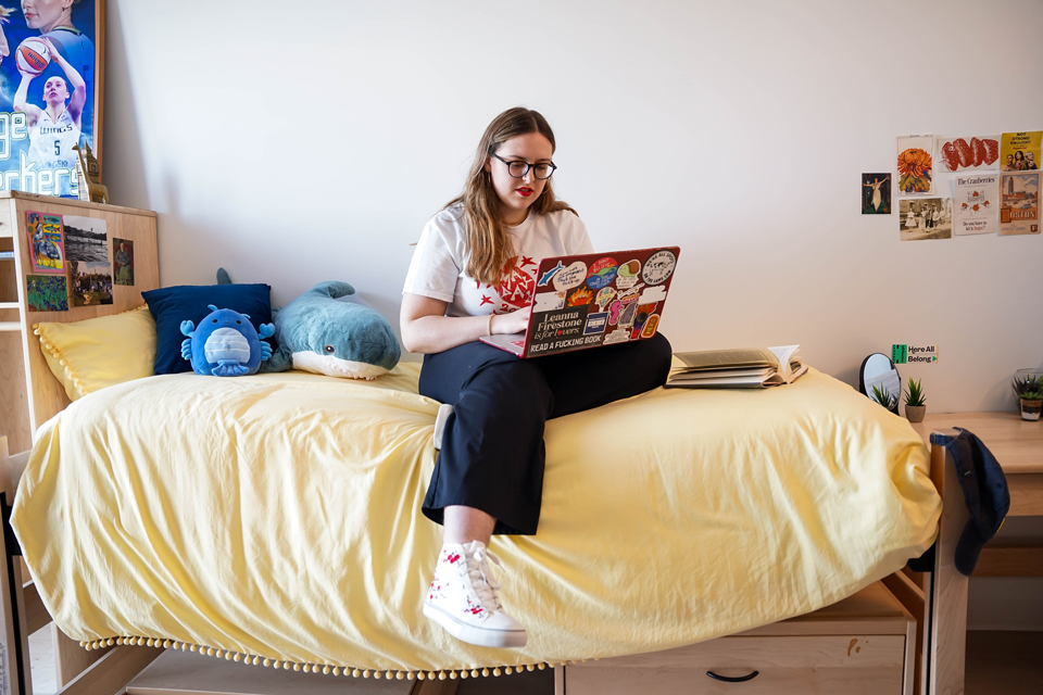 A Simmons student sitting on a bed in one of the student rooms in the new Living-Learning Center at Simmons University
