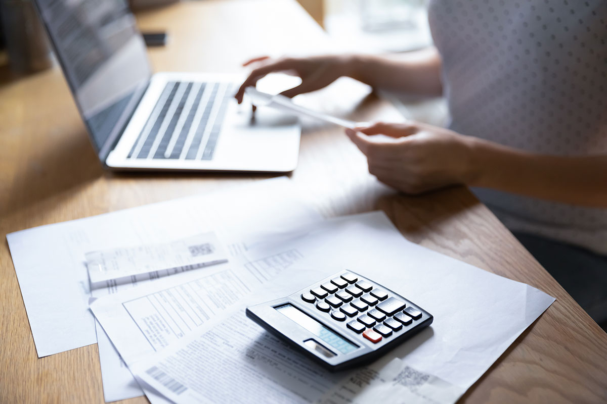 A close-up photo of a persons hand on a laptop keyboard. Nearby on the same desk is a calculator on top of some papers.