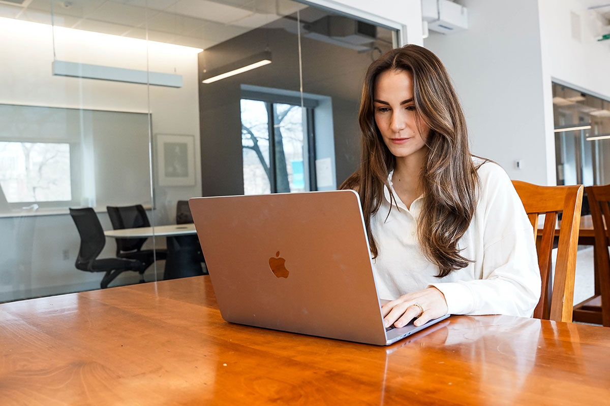 A Simmons student working on a laptop while seated at a wooden table