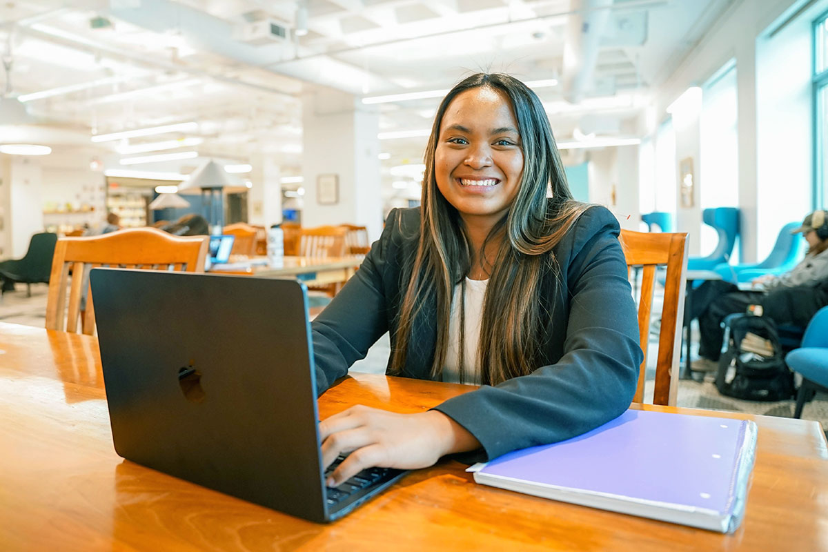 A Simmons student wearing a blazer while working on a laptop seated at a table in a library