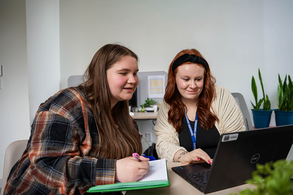 Two students working together taking notes and looking at laptop