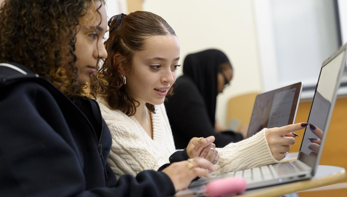 Two students in classroom desks looking at one laptop screen