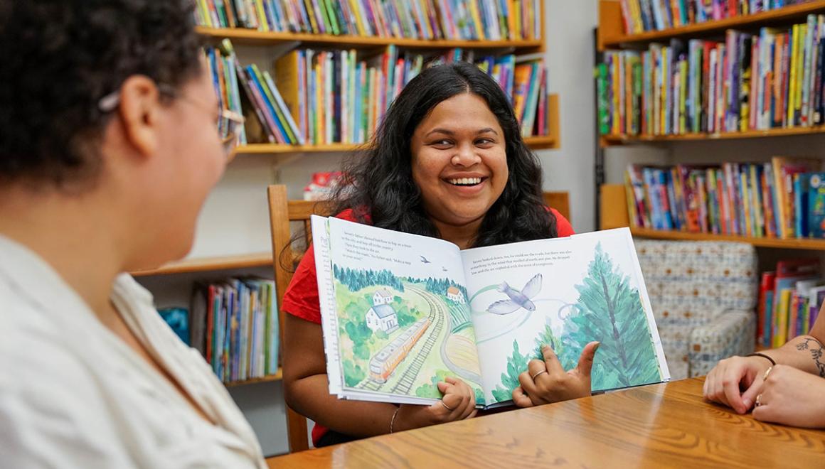 A Simmons Children's Literature student holds an open picturebook while seated at a table in a children's library