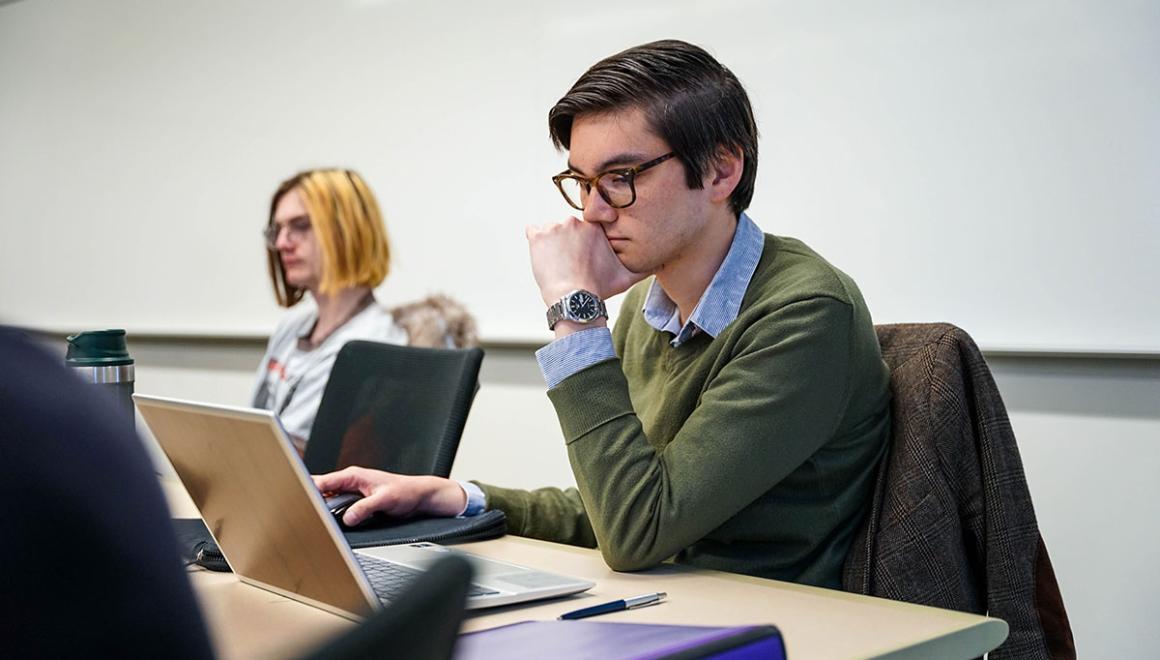 A graduate student seated at a desk looking thoughtful while looking at an open laptop