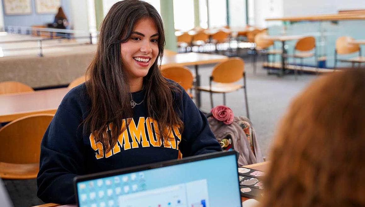 Two Simmons Behavior Analysis students talking across a desk. One has a laptop open.