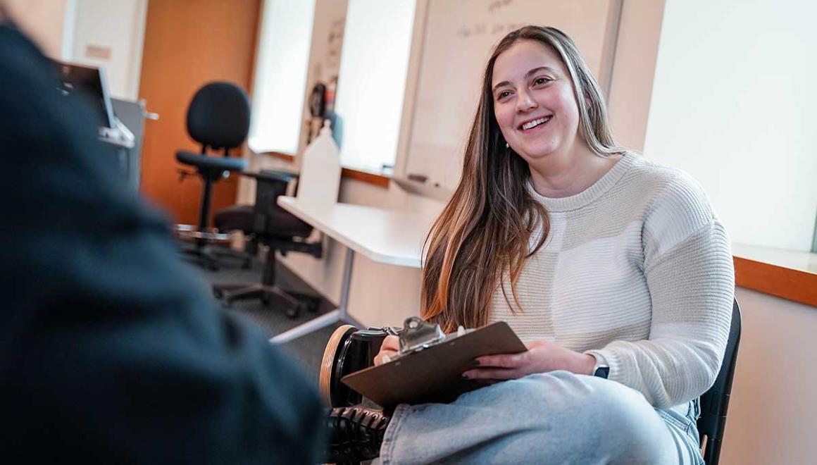 A social work student sitting and talking with another student while holding a clipboard