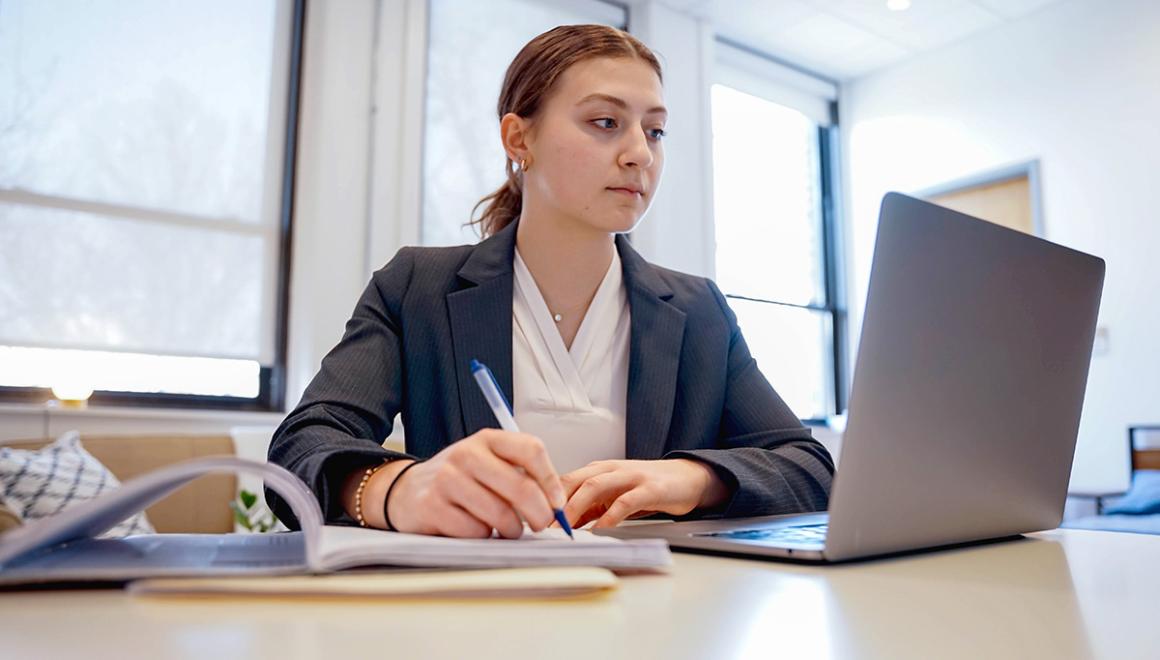 A Simmons management student taking notes while looking at a laptop.