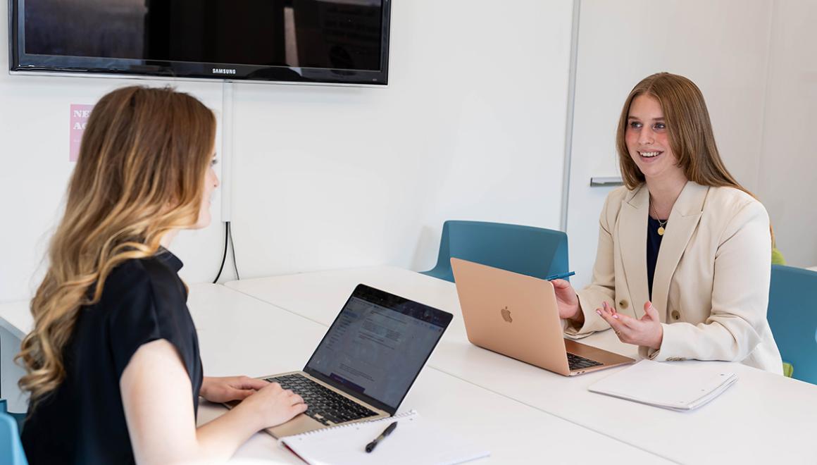 Two Simmons management students working on laptops at a table in a conference room