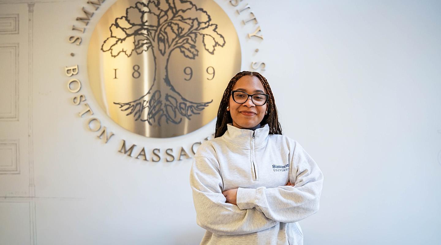 Student standing in front of Simmons University emblem. Smiling with arms crossed.