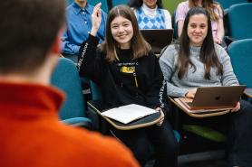 Two Simmons undergraduate students in a class. One of them is raising their hand.