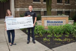 Two staff members stand in front of the Simmons sign holding a big check