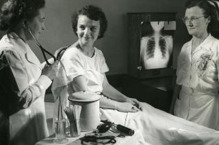 Dr. Marjorie Readdy, Director of Health Services, 1949-1982, poses with a stethoscope on the back of an unidentified nursing student while Elsie Fenney, Assistant Director of Health Services, looks on.