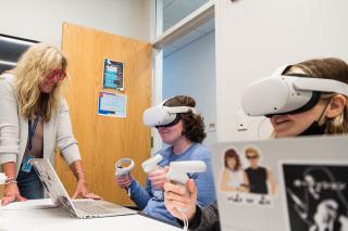 Students wearing VR headsets and holding controllers sitting in front of laptops with a faculty member assisting.