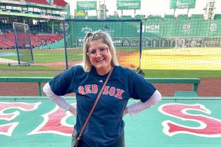 Photo of Emily Mills standing by the Red Sox dugout at Fenway Park with a view of the field behind her