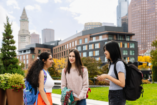 Three Simmons students talking in the Academic quad, with a view of some of Boston's tall buildings in the background