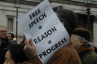 A protestor holds a sign that reads "Free Speech = Reason = Progress"