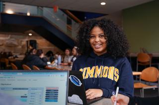 Student in Simmons sweatshirt smiling while on laptop in the Fens