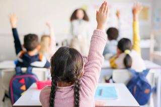 View from the back of an elementary school classroom where students are raising their hands