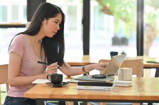 An online student sitting at a table near windows and pointing to a laptop screen on a desk