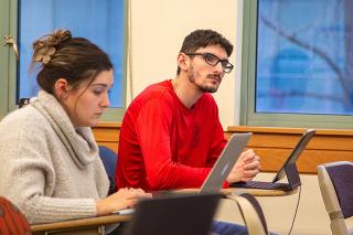 Two students in classroom at desks. One looking down at a laptop - the other has an tablet with a keyboard and is looking forward.