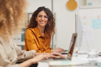 Two students working at a long desk. One is wearing an orange shirt and is smiling at the other.