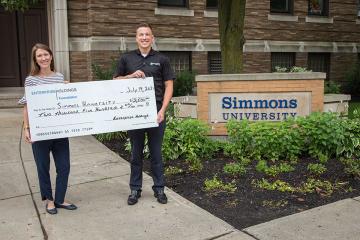 Two staff members stand in front of the Simmons sign holding a big check