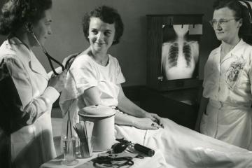 Dr. Marjorie Readdy, Director of Health Services, 1949-1982, poses with a stethoscope on the back of an unidentified nursing student while Elsie Fenney, Assistant Director of Health Services, looks on.