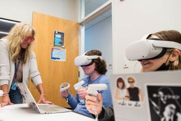 Students wearing VR headsets and holding controllers sitting in front of laptops with a faculty member assisting.