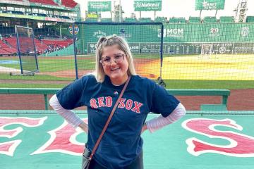 Photo of Emily Mills standing by the Red Sox dugout at Fenway Park with a view of the field behind her