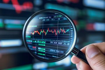 A hand holds a magnifying glass, focusing on a volatile stock chart displayed on a computer screen