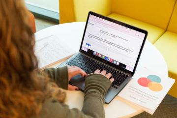 A student at a round table using a laptop with an open notebook on the table