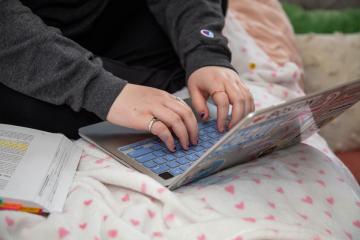 Closeup photo of a Simmons student working on a laptop on their bed in their residence hall room