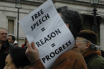 A protestor holds a sign that reads "Free Speech = Reason = Progress"