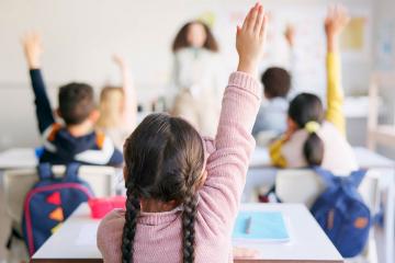 View from the back of an elementary school classroom where students are raising their hands
