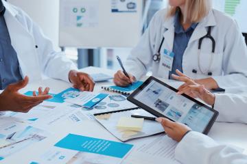 A number of healthcare workers at a table covered with pamphlets and papers.  One of them is using a tablet.