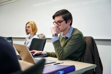 A graduate student seated at a desk looking thoughtful while looking at an open laptop
