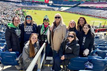 A group of Simmons students with Dean Astrid Sheil and others wearing "Study Women's Sports" shirts at a Legacy soccer game.