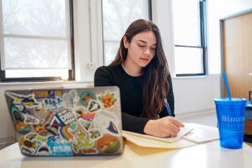 A Simmons management student working at a desk with an open laptop.  A blue Simmons cup is on the table.