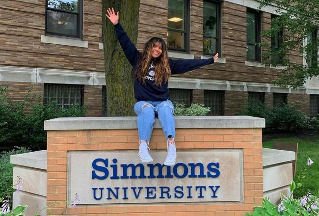Simmons University student sitting on the Simmons University sign in Boston, Massachusetts.