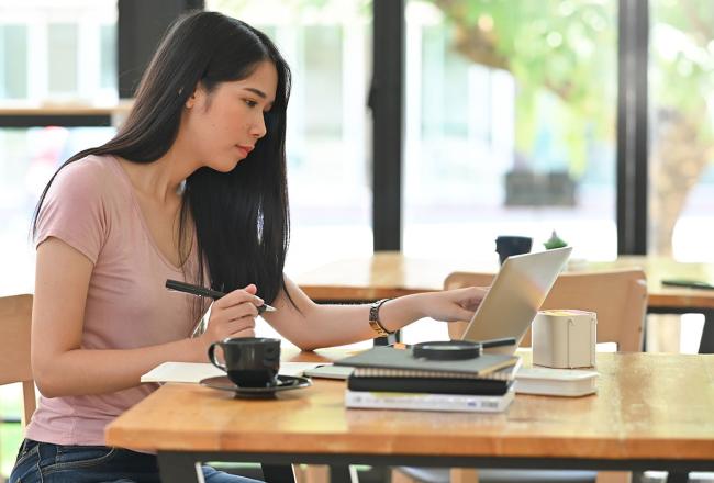 An online student sitting at a table near windows and pointing to a laptop screen on a desk