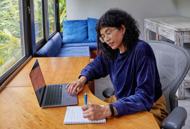 An adult woman wearing a blue shirt while working on a laptop and writing on a pad