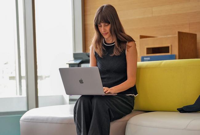 A Simmons student sitting on a yellow couch while working on a laptop