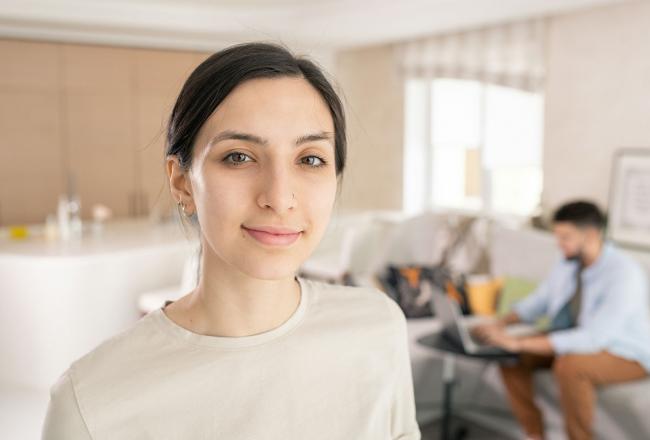 A young woman smiling in the foreground while a man sits on a couch working on a laptop in the background.  Photo courtesy of unsplash.