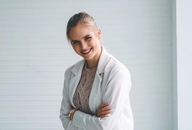 A young woman wearing a white lab coard and standing near a blank wall. Courtesy of unsplash