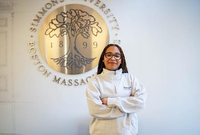 Student standing in front of Simmons University emblem. Smiling with arms crossed.