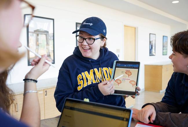 A student wearing a Simmons sweatshirt holding a tablet and pointing to the screen
